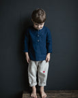 A young boy wearing a blue shirt and gray pants stands on a wooden bench against a dark background.