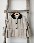 A beige jacket with a black collar is hanging on a wooden ladder against a white wall.