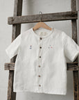 A white button-down shirt with a small anchor embroidered on the left side hangs on a wooden ladder against a white wall.