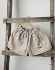 A pair of beige shorts with a small embroidered chicken design is hanging on a wooden ladder against a white wall.