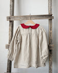 A beige dress with a red collar is hanging on a wooden ladder against a white wall.