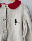 A close-up view of a beige blouse with a red collar and a black cat embroidered on the front.