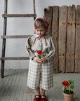 A young girl in a checkered dress and red shoes holds a red flower in front of a wooden ladder and a potted plant.
