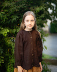 A young girl with long blonde hair stands in front of a lush green bush, wearing a brown blouse with puffed sleeves and a brown skirt.