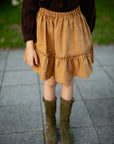 A person wearing a brown top, a yellow skirt, and green boots stands on a tiled surface.