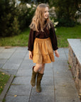 A young girl with long blonde hair is walking on a sidewalk, wearing a brown sweater, a yellow skirt, and green boots.