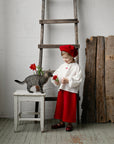 A young girl in a red hat and white blouse stands next to a gray cat, holding a flower, with a wooden ladder and a wooden fence in the background.