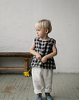 A young child in a black and white checkered shirt and white pants stands in a room, holding a yellow flower in their hand.