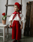 A young child dressed in a red outfit and hat stands in a room with a wooden ladder and a potted plant.
