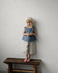 A young girl with blonde hair is standing on a wooden bench, wearing a blue top and plaid shorts, and holding a small object in her hands.