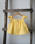 A yellow dress with ruffled sleeves is hanging on a wooden ladder against a white wall.