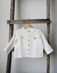 A white button-down shirt with a small embroidered animal design on the left side is hanging on a wooden ladder against a white wall.