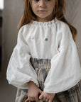 A young girl with long brown hair wearing a white blouse with embroidered flowers and a gray skirt, holding a basket of white flowers.