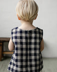 A young child with blonde hair is standing in front of a plain white wall, wearing a blue and white checkered shirt.