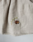 a close-up view of a beige fabric with a small embroidered design featuring a pine cone and a small animal, likely a hedgehog, on it.