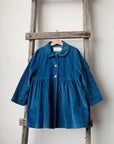 A blue dress with a ruffled collar and buttons is hanging on a wooden ladder against a white wall.