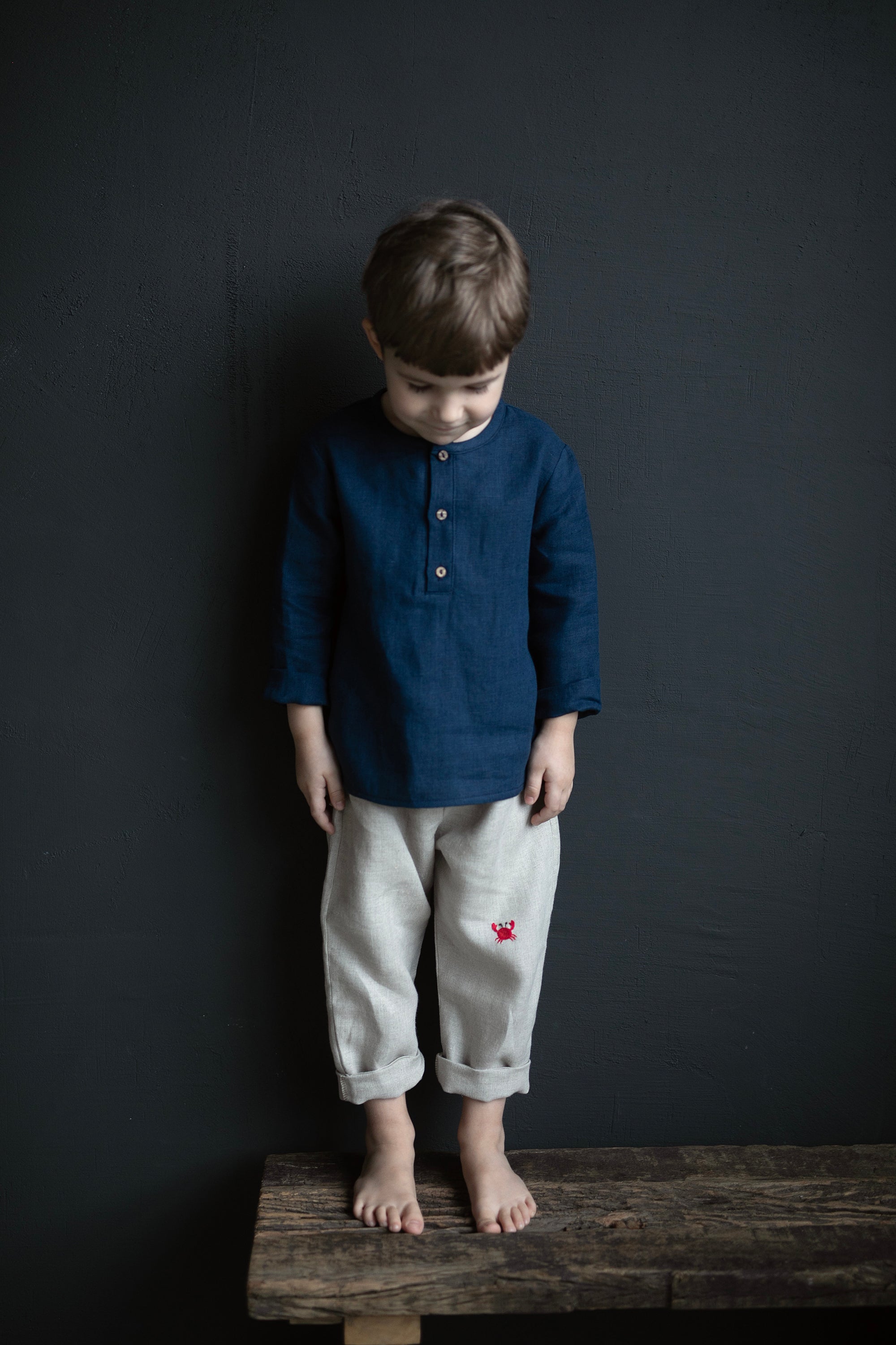 A young boy wearing a blue shirt and gray pants stands on a wooden bench against a dark background.