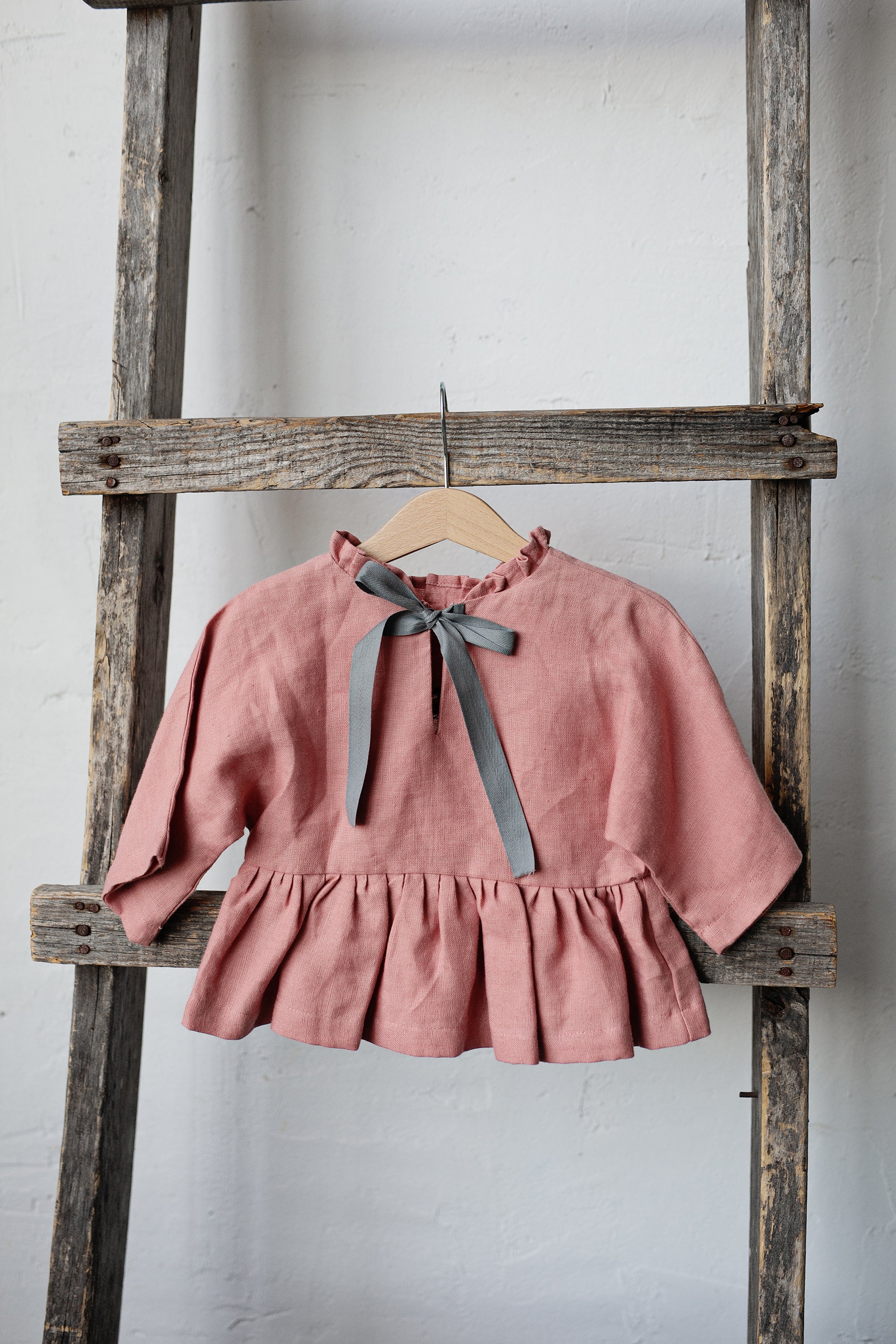 A pink ruffled blouse with a gray ribbon tied in a bow is hanging on a wooden ladder against a white wall.