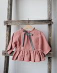 A pink ruffled blouse with a gray ribbon tied in a bow is hanging on a wooden ladder against a white wall.