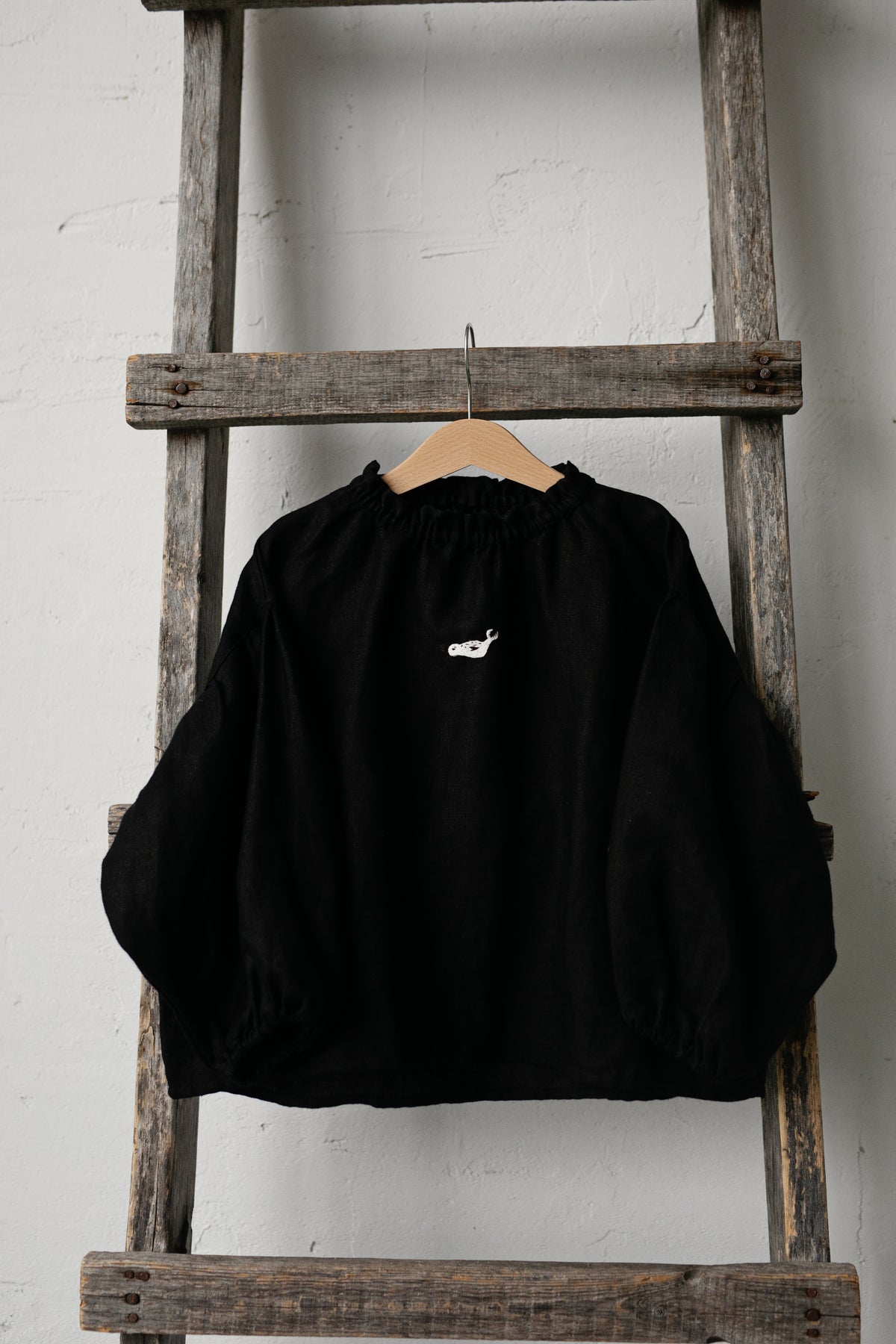 A black sweater with a white logo is hanging on a wooden ladder against a white wall.