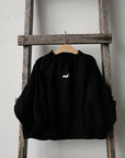 A black sweater with a white logo is hanging on a wooden ladder against a white wall.