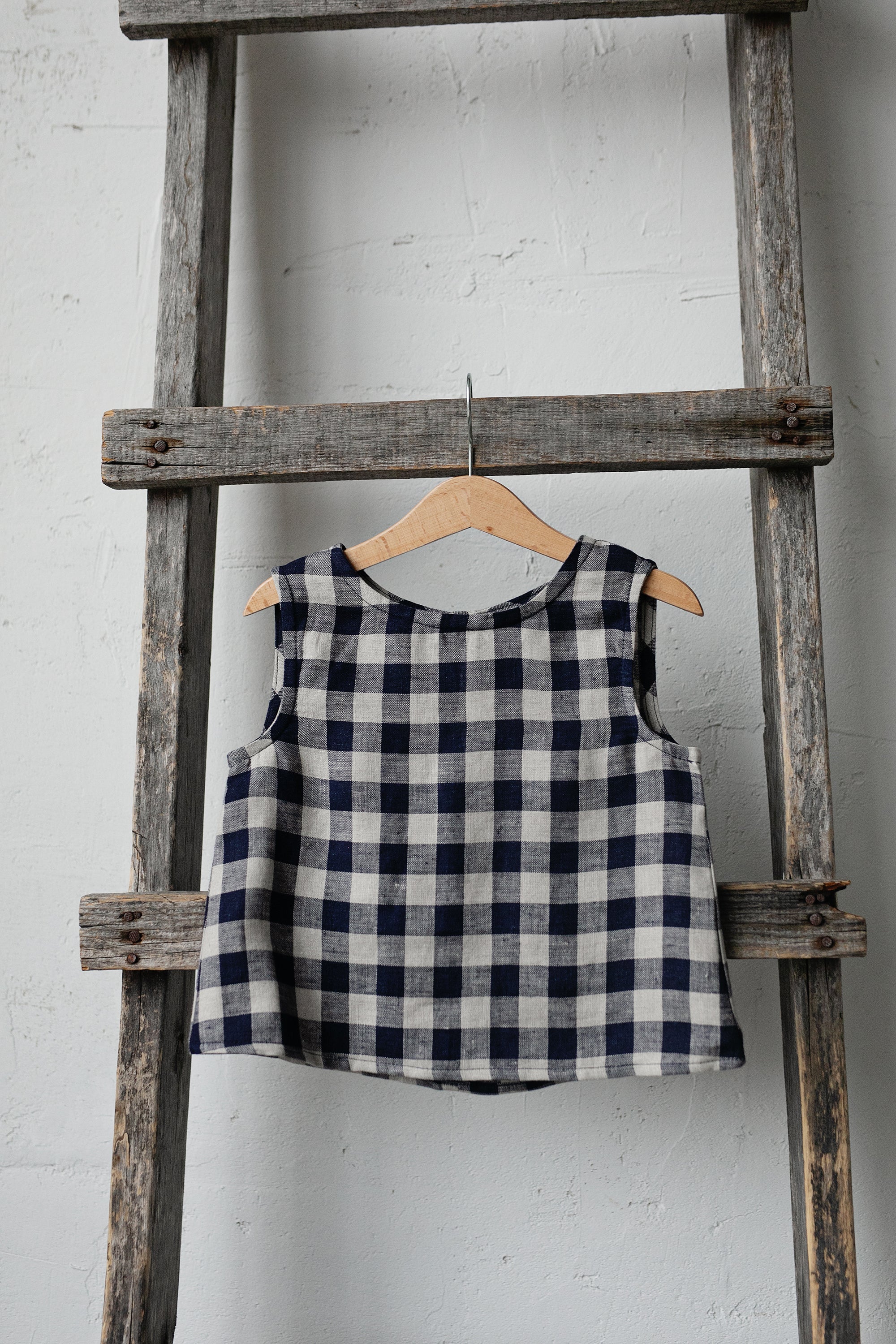 A blue and white checkered top is hanging on a wooden ladder against a white wall.