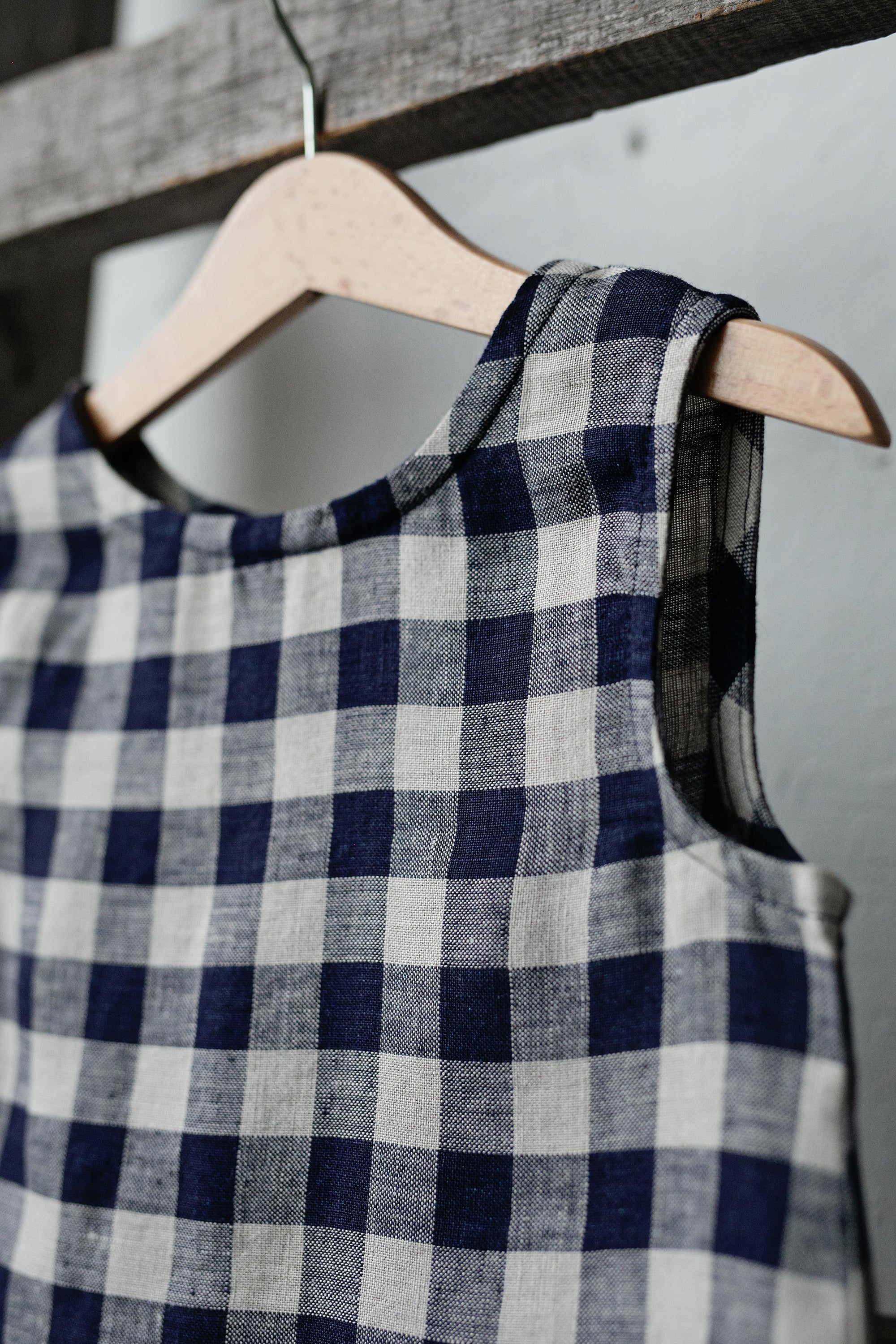 A blue and white checkered dress is hanging on a wooden hanger against a white wall.