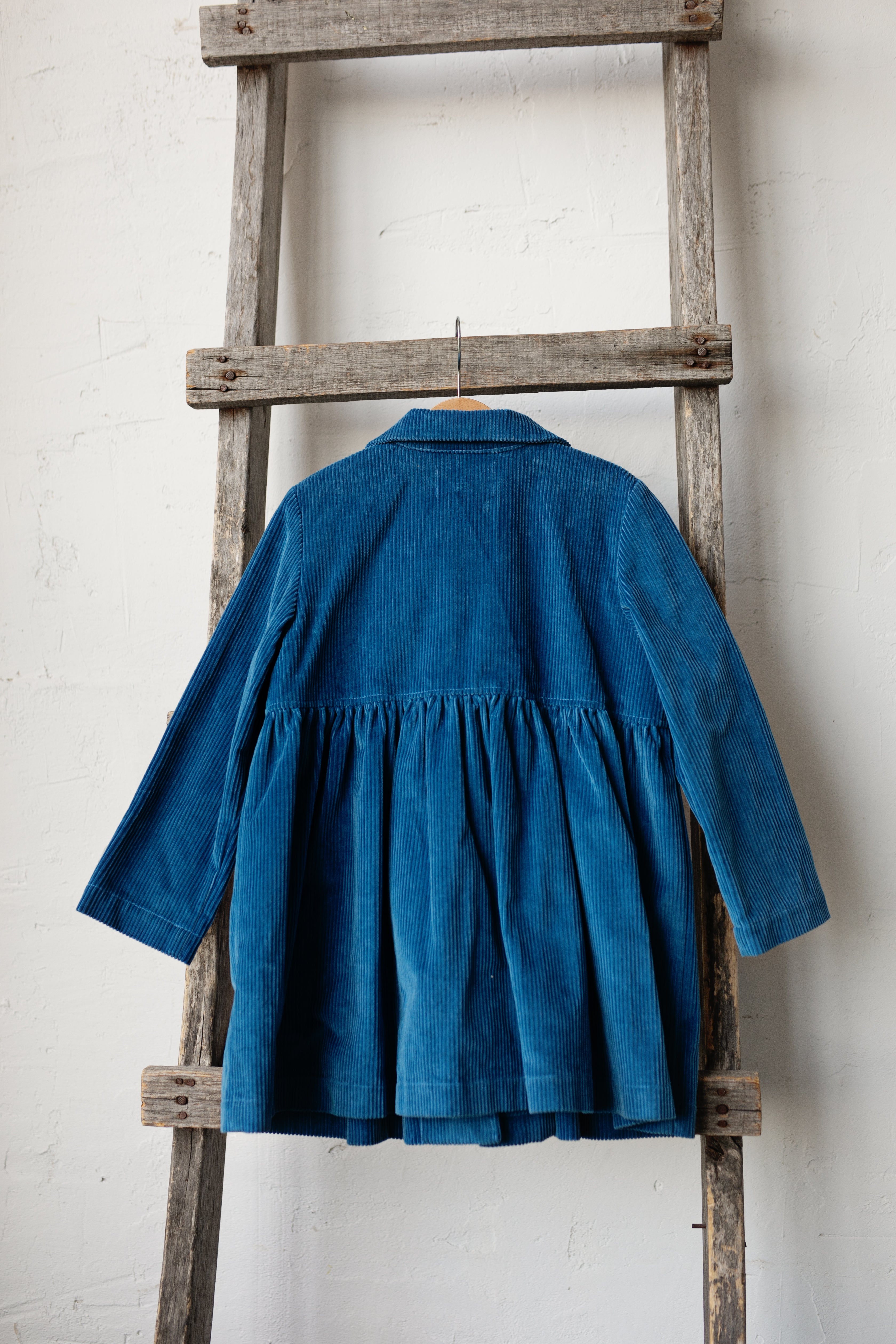 A blue corduroy dress is hanging on a wooden ladder against a white wall.