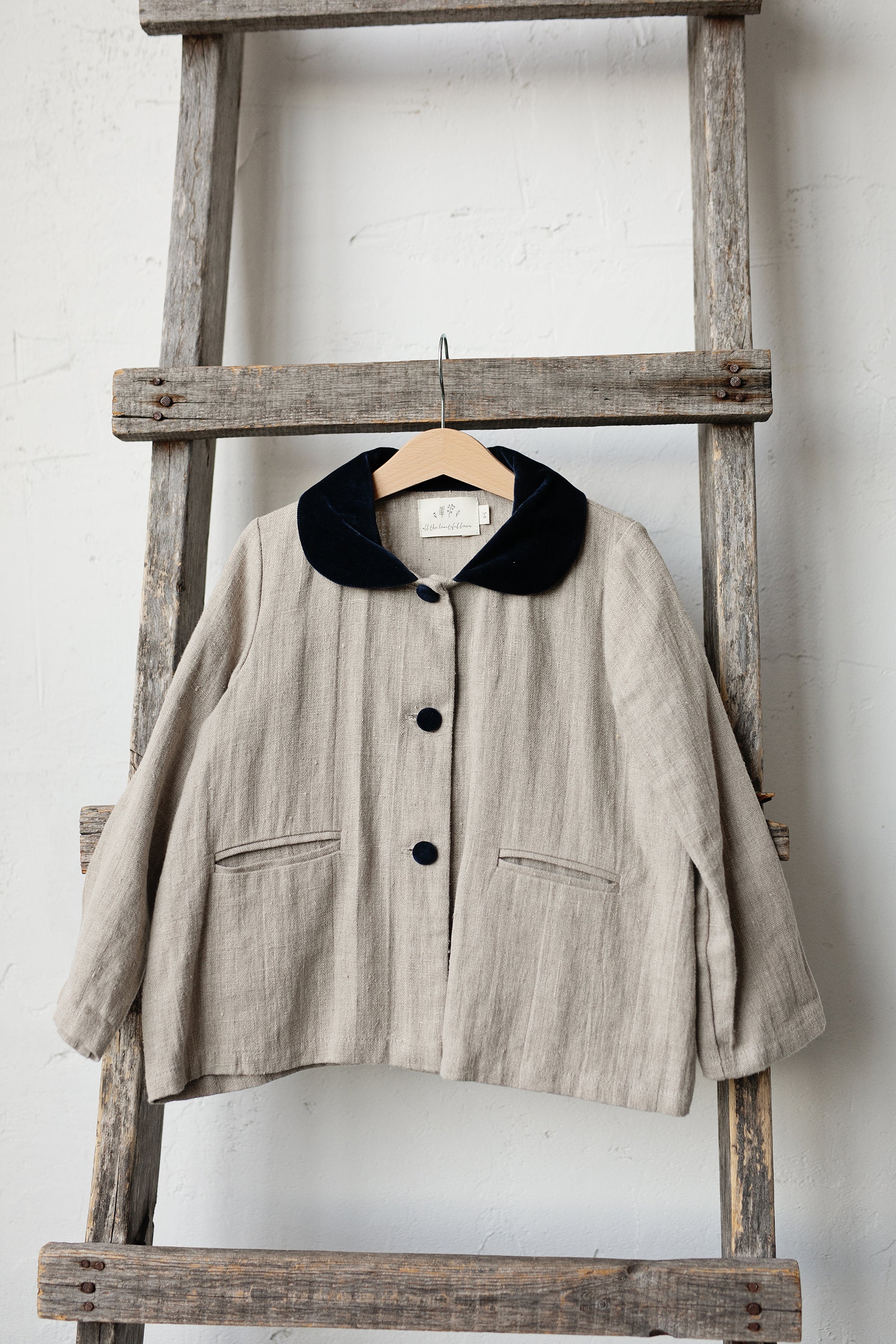 A beige jacket with a black collar is hanging on a wooden ladder against a white wall.