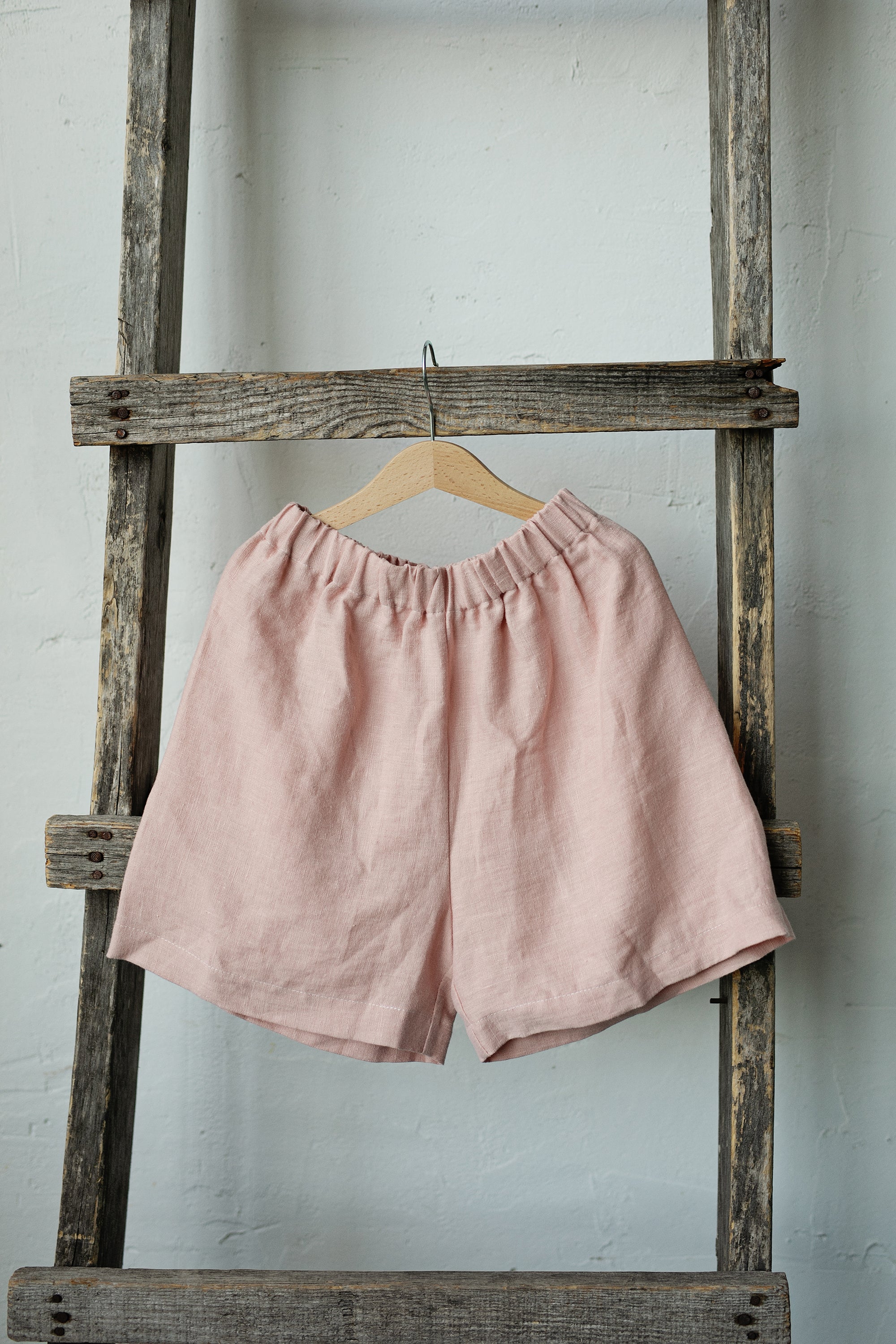 A pair of pink shorts is hanging on a wooden ladder against a white wall.