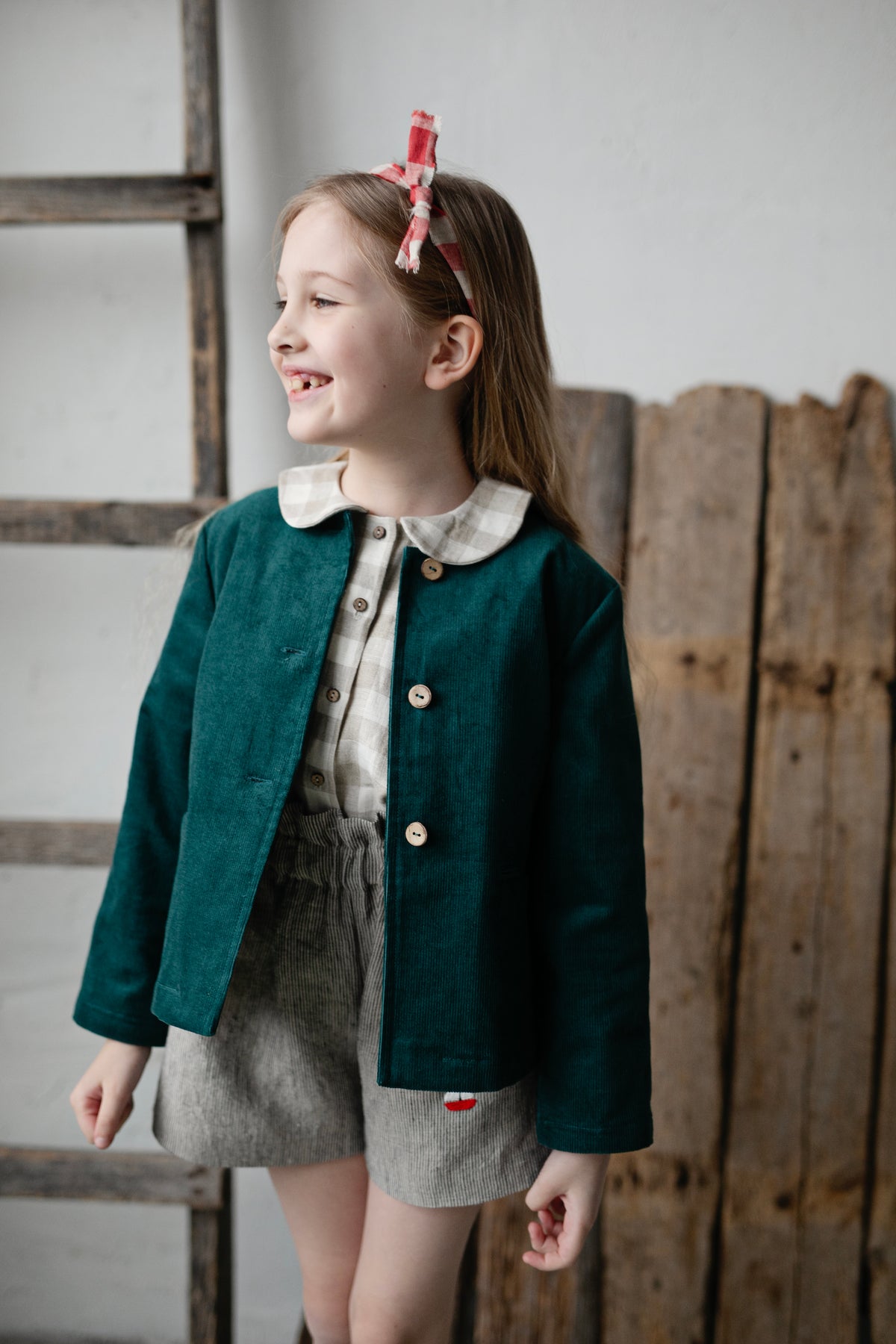A young girl with a green jacket and gray shorts is smiling and looking to the side, with a wooden fence and ladder in the background.