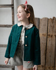 A young girl with a green jacket and gray shorts is smiling and looking to the side, with a wooden fence and ladder in the background.