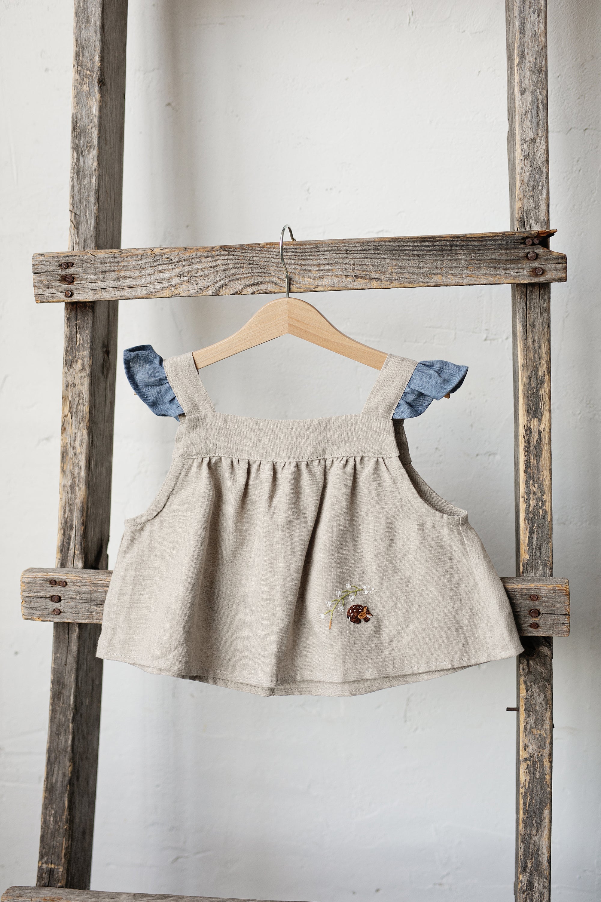 A beige dress with blue accents hangs on a wooden ladder against a white wall.