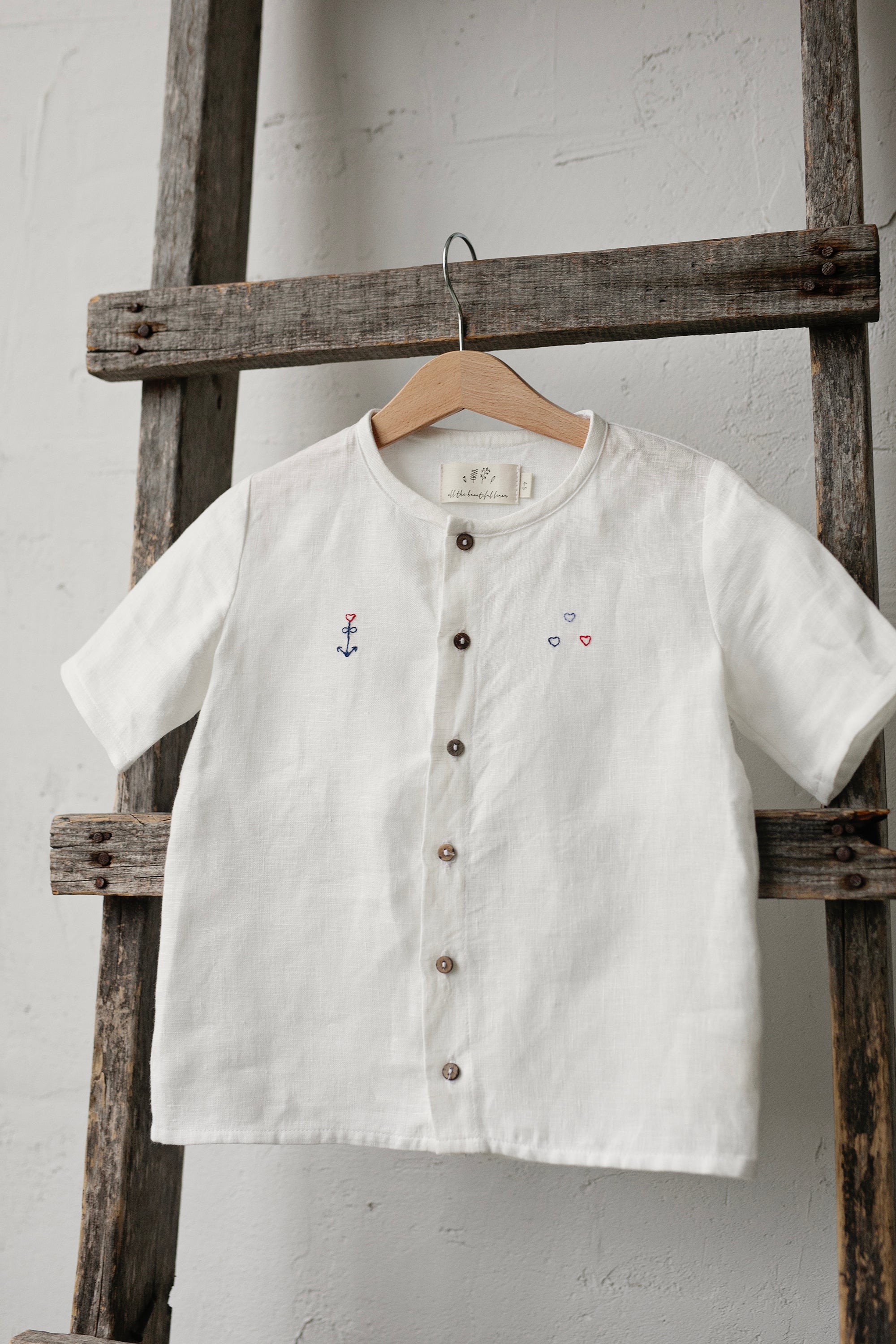 A white button-down shirt with a small anchor embroidered on the left side hangs on a wooden ladder against a white wall.