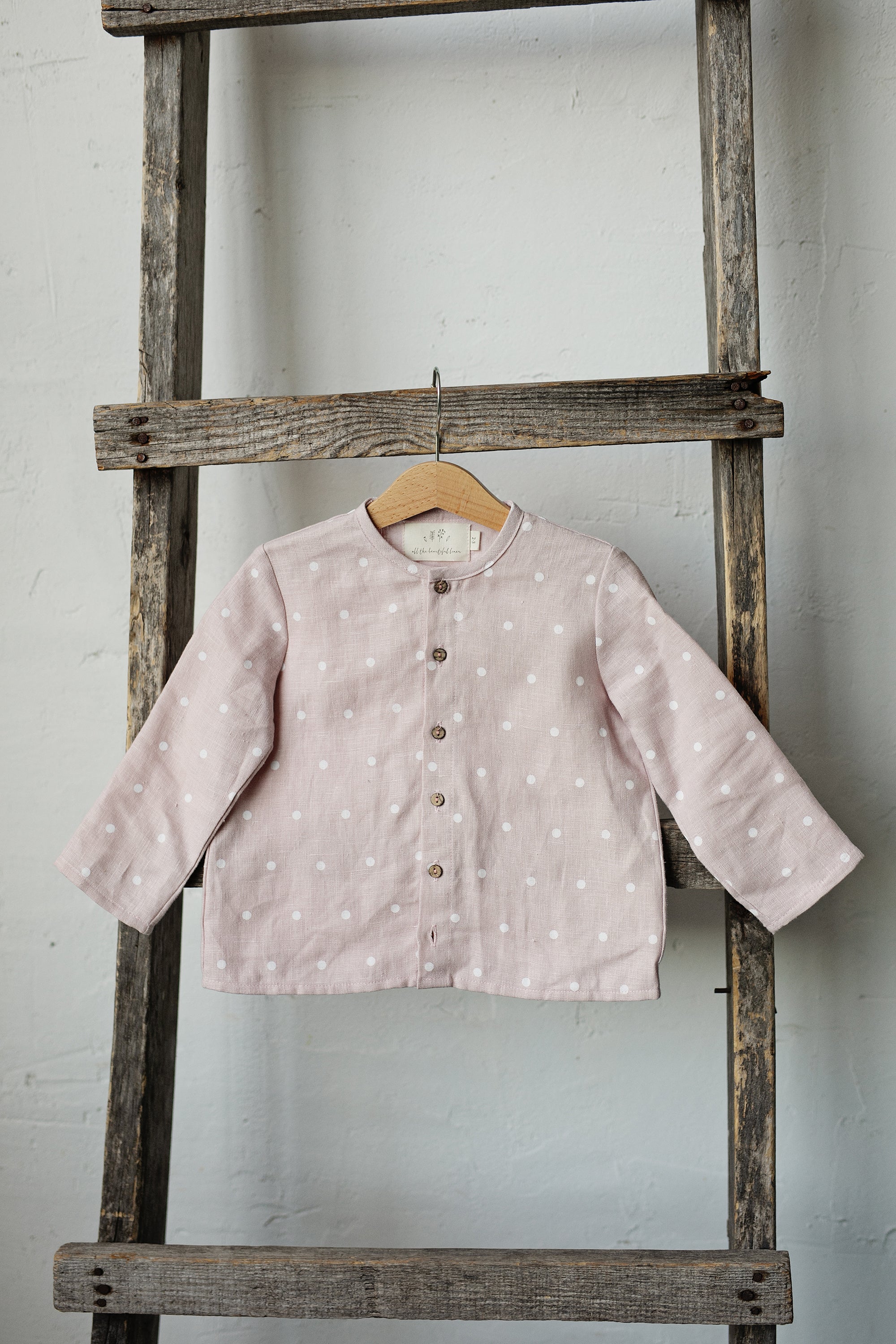 A pink polka dot shirt is hanging on a wooden ladder against a white wall.