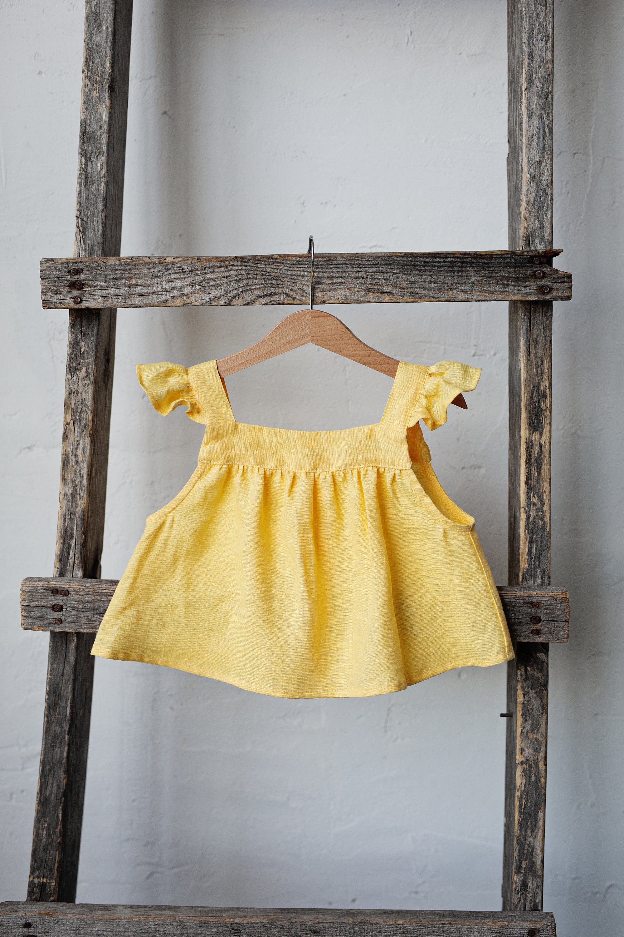 A yellow dress with ruffled sleeves is hanging on a wooden ladder against a white wall.