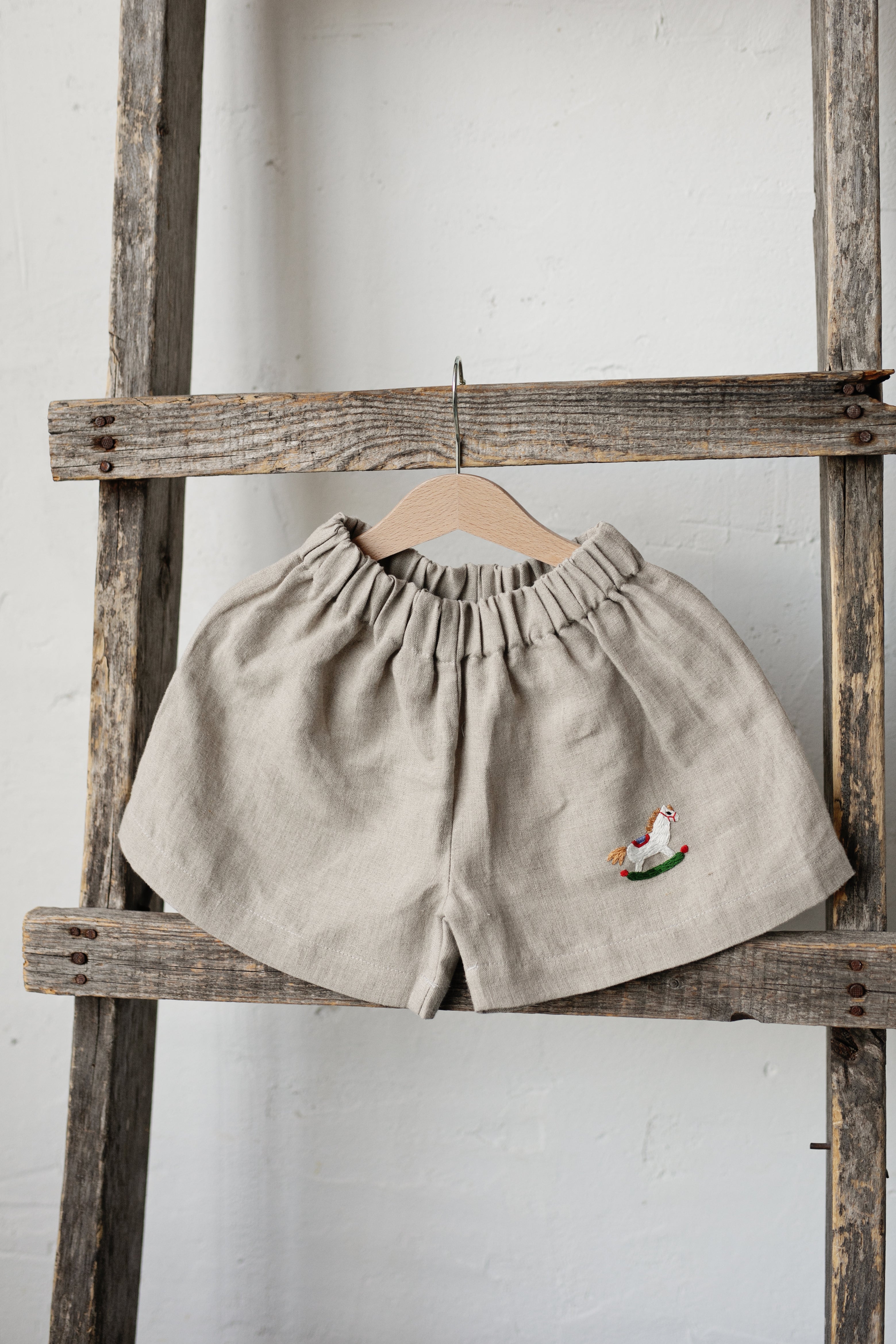 A pair of beige shorts with a small embroidered chicken design is hanging on a wooden ladder against a white wall.