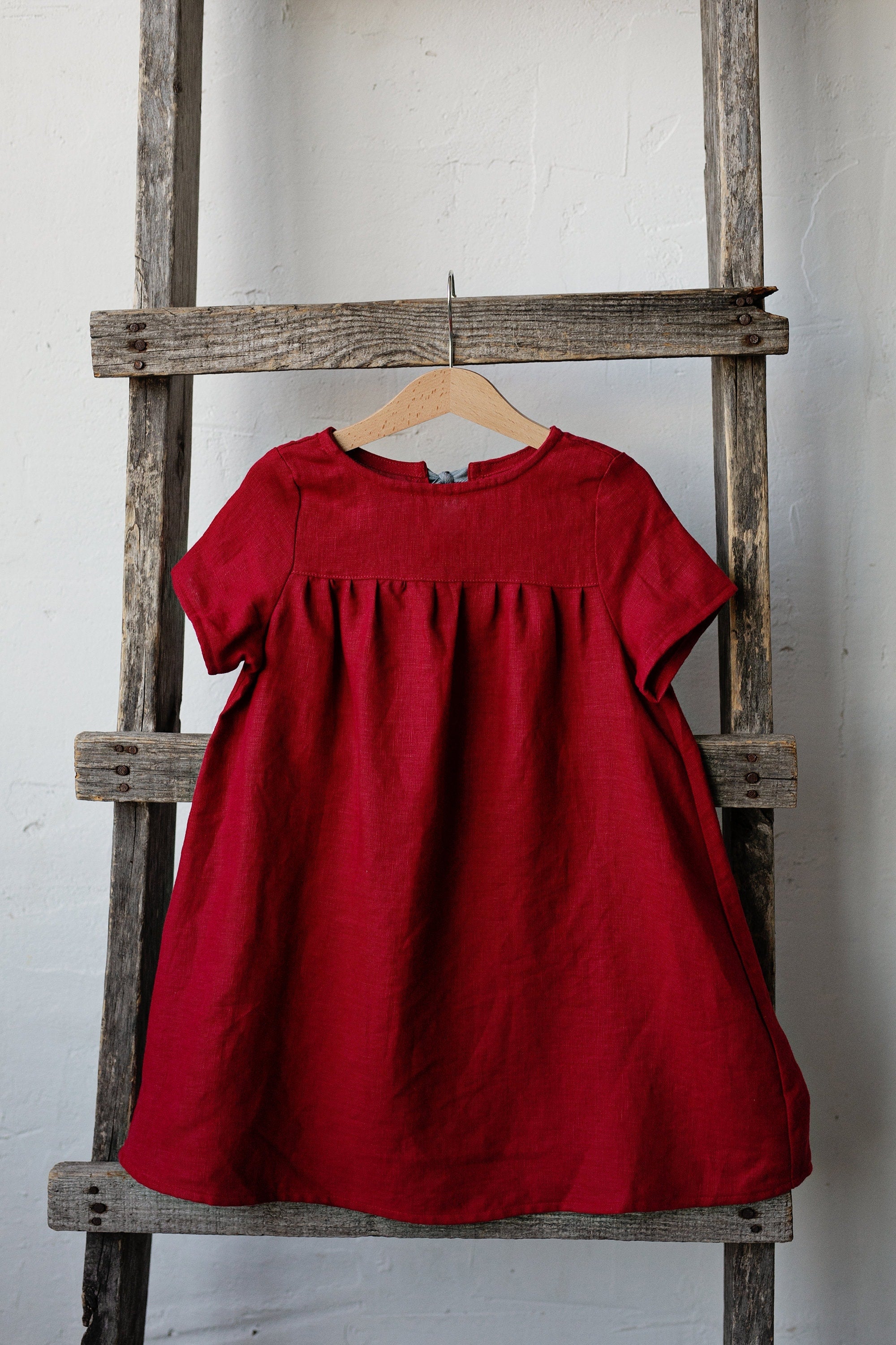 A red dress is hanging on a wooden ladder against a white wall.