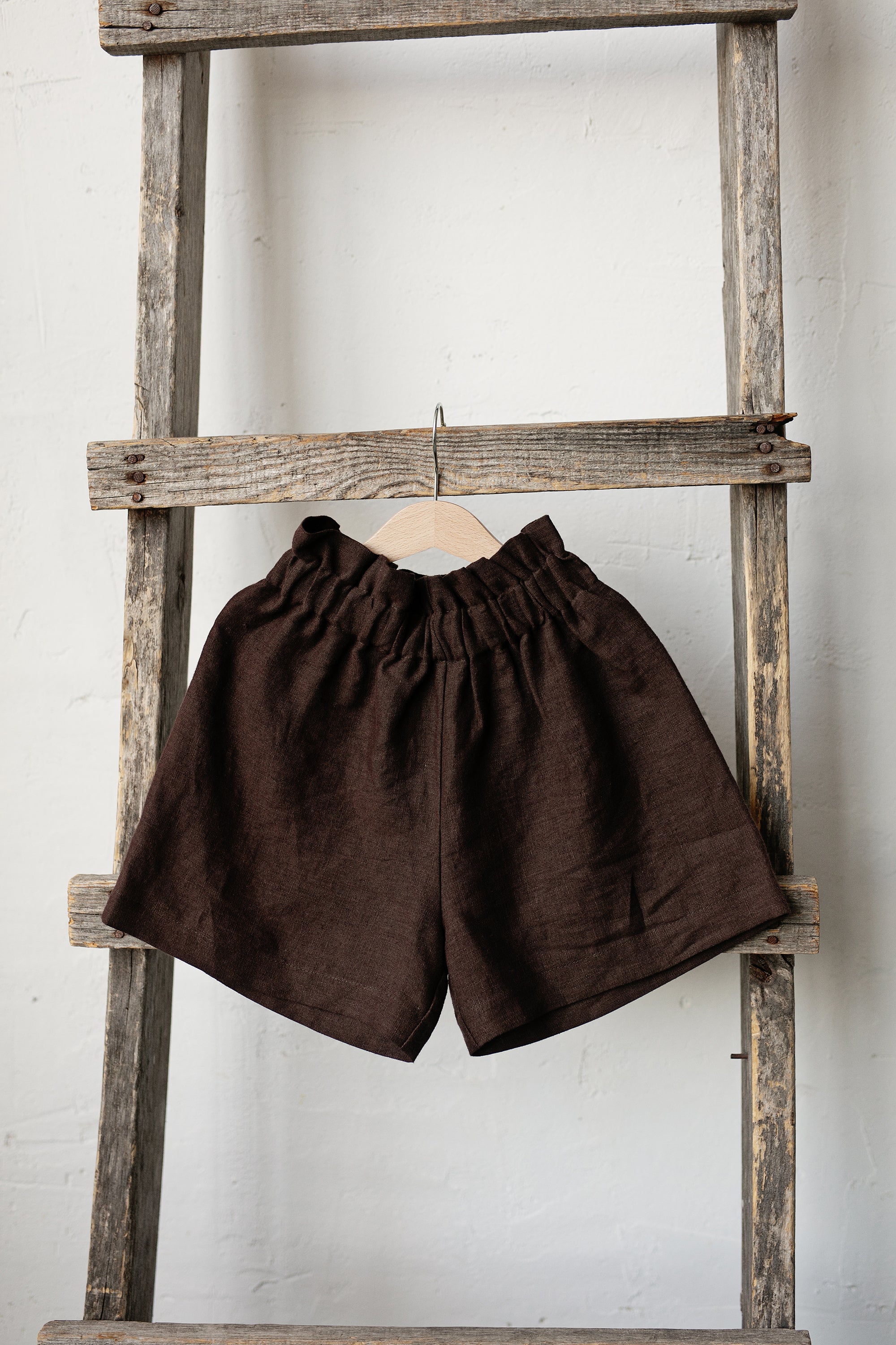 A pair of brown shorts hanging on a wooden ladder against a white wall.