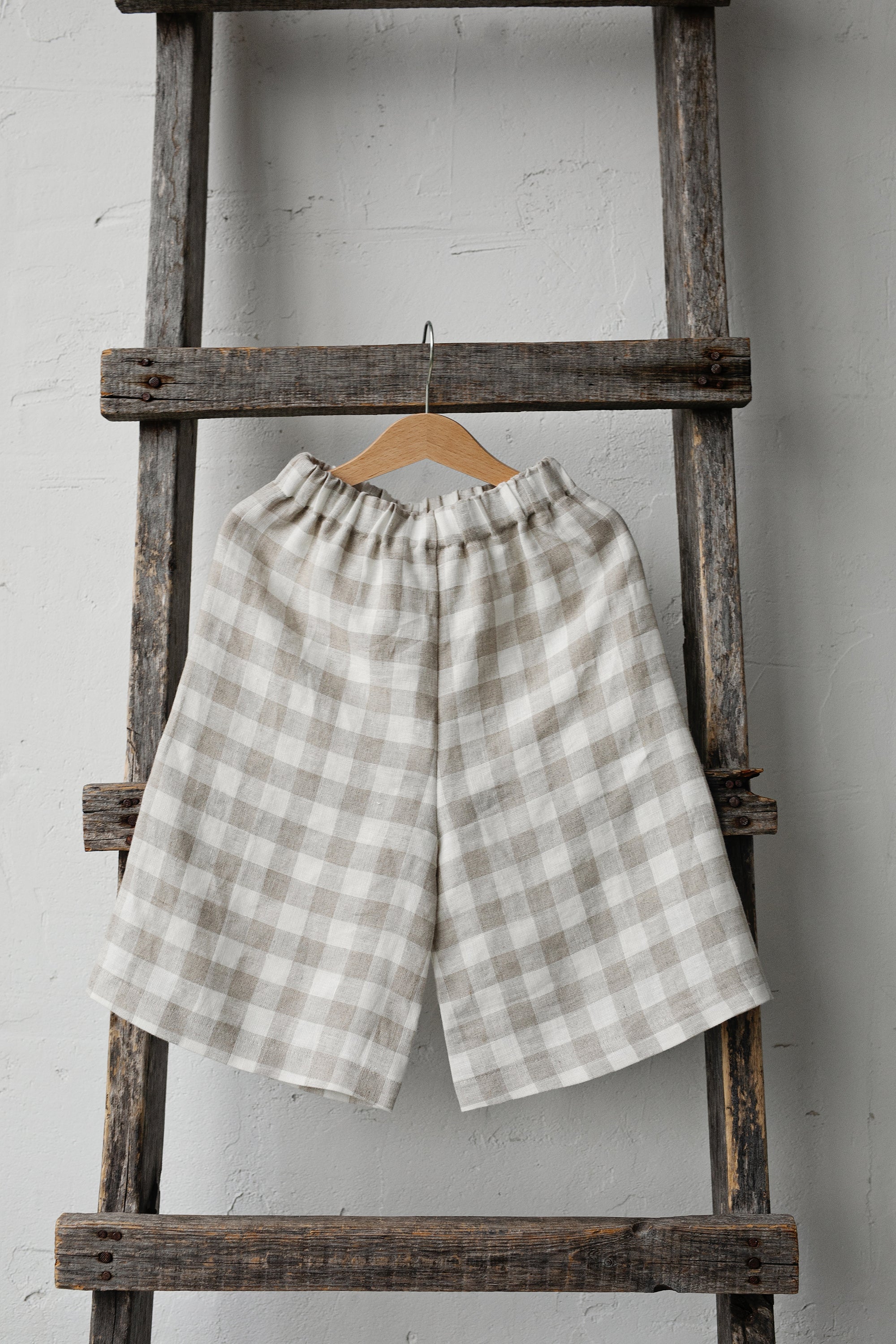 A pair of gray and white checkered shorts is hanging on a wooden ladder against a white wall.
