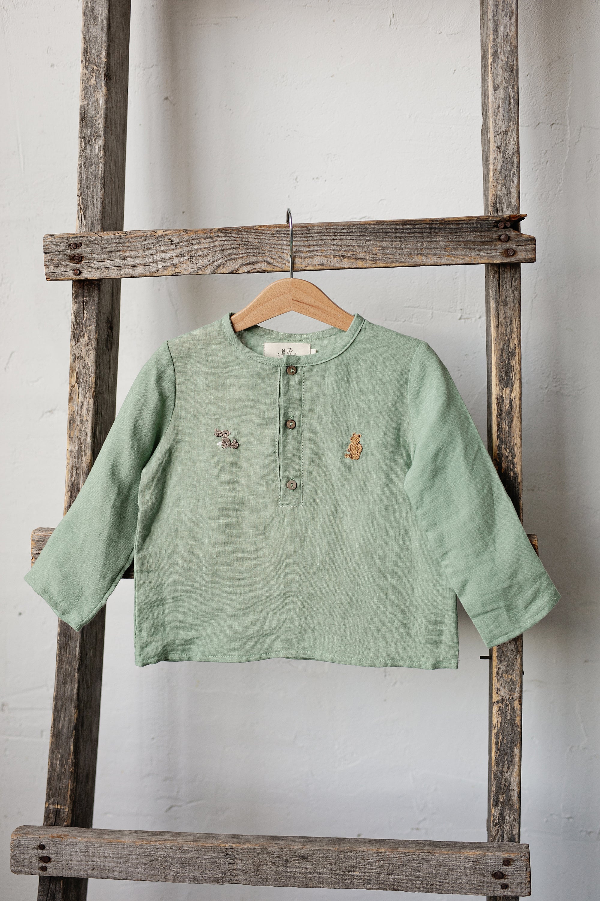 A light green button-down shirt is hanging on a wooden ladder against a white wall.