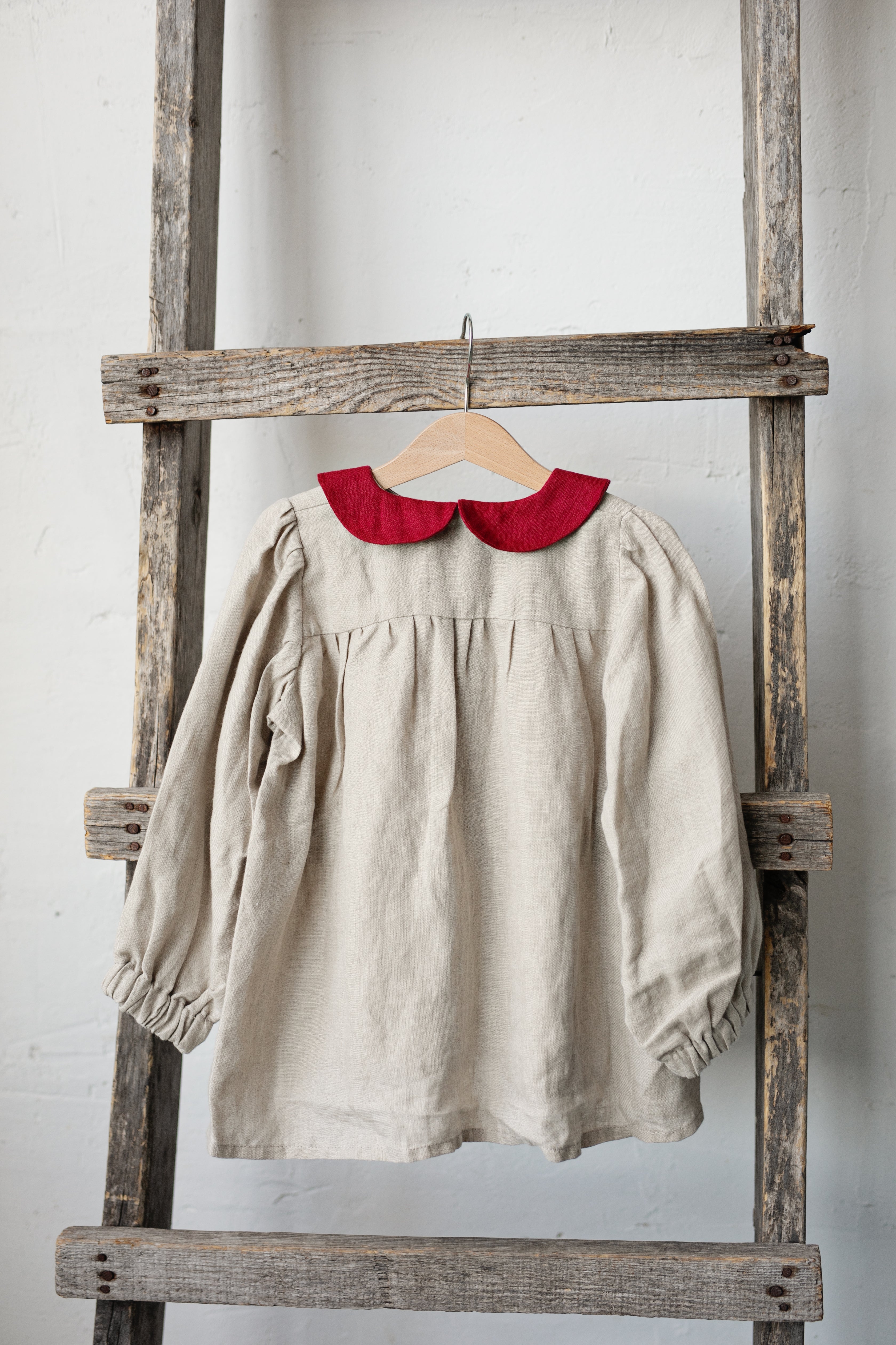 A beige dress with a red collar is hanging on a wooden ladder against a white wall.