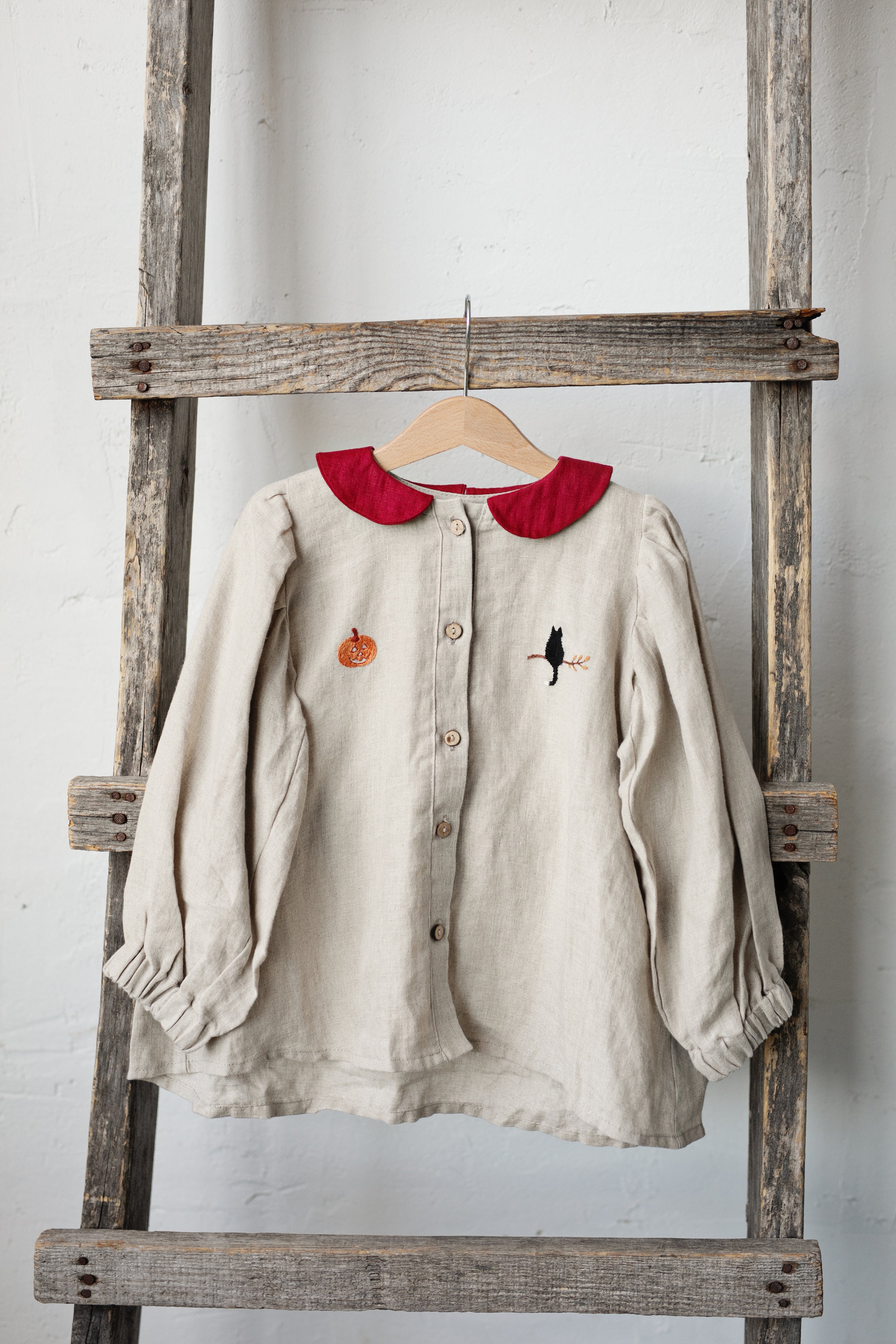 A beige blouse with a red collar and a small black bird embroidered on the front is hanging on a wooden ladder against a white wall.
