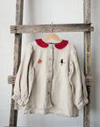 A beige blouse with a red collar and a small black bird embroidered on the front is hanging on a wooden ladder against a white wall.