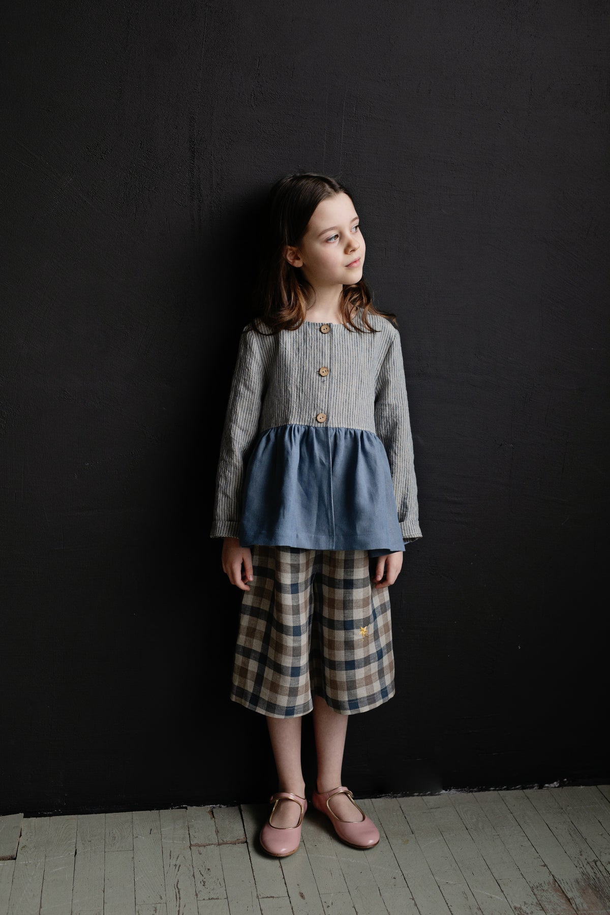 A young girl in a gray and blue dress stands against a black background, looking off to the side with a thoughtful expression.