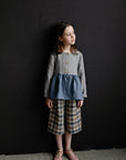 A young girl in a gray and blue dress stands against a black background, looking off to the side with a thoughtful expression.
