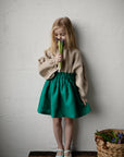 A young girl in a green skirt and beige blouse stands next to a basket of flowers, holding a bunch of flowers in her hand.