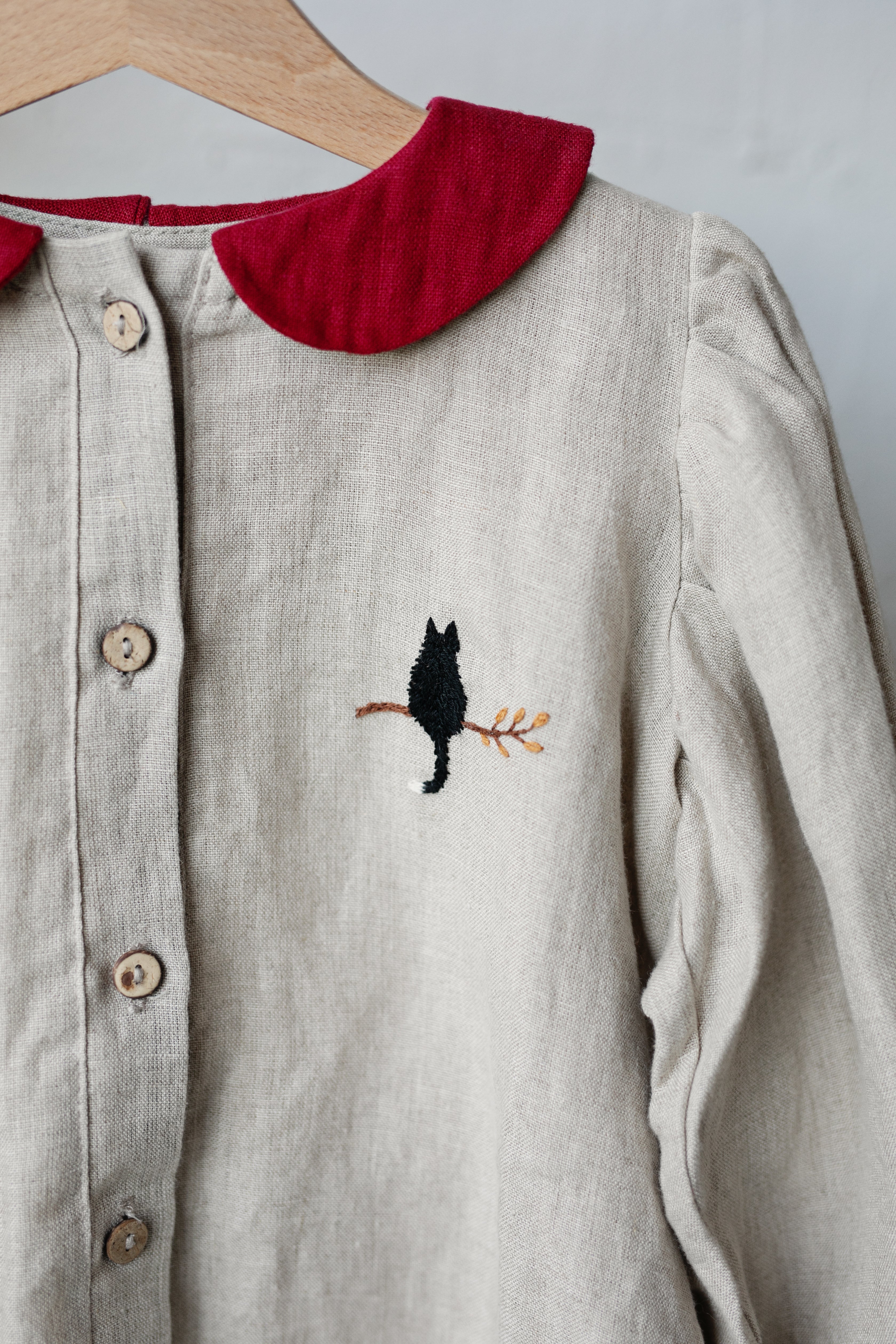 A close-up view of a beige blouse with a red collar and a black cat embroidered on the front.