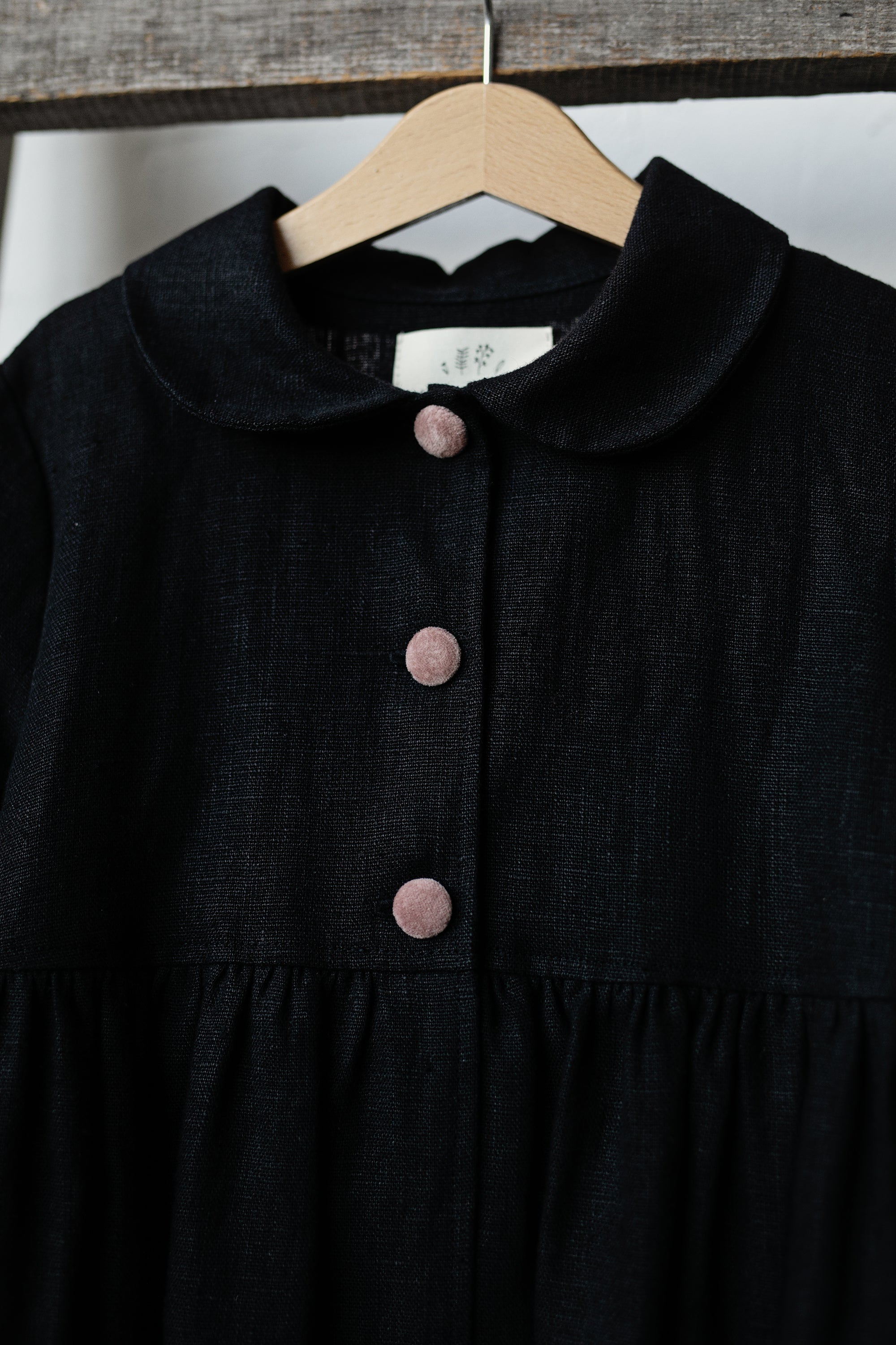 A black dress with a ruffled collar and three buttons, hanging on a wooden hanger.