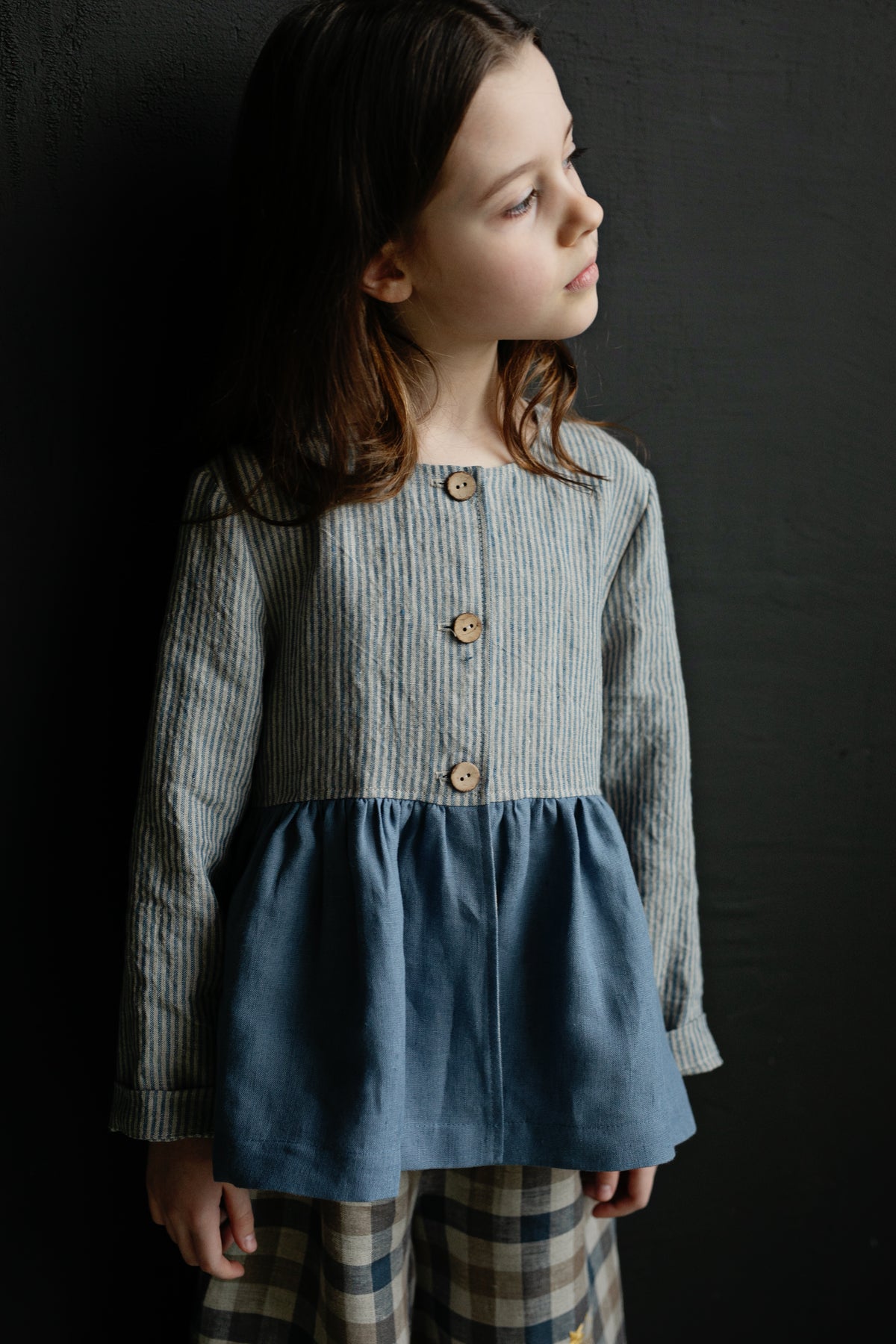 A young girl with long brown hair wearing a striped jacket and plaid pants stands against a dark background.